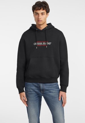 Svart bomullssweatshirt med hette, frontlomma og brodert "GUESS JEANS"-logo i rødt og hvitt. Brukt sammen med lyseblå jeans.