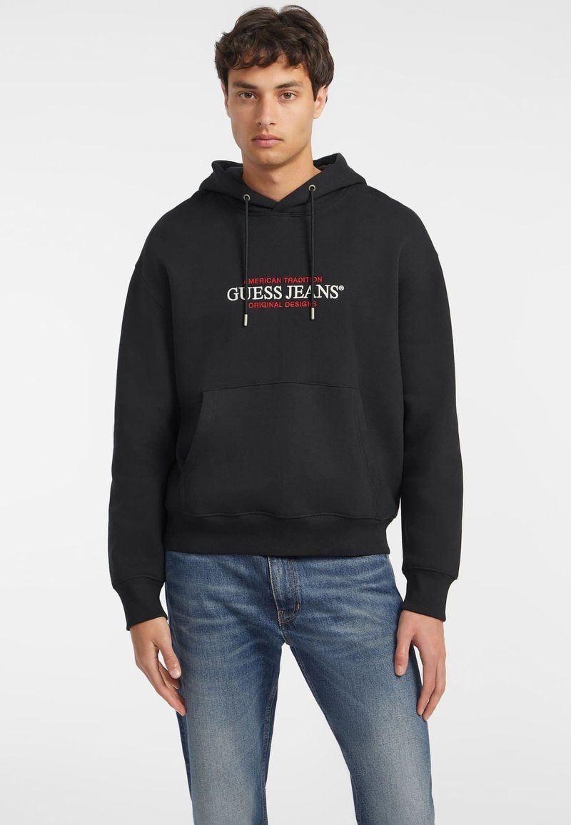 Svart bomullssweatshirt med hette, frontlomma og brodert "GUESS JEANS"-logo i rødt og hvitt. Brukt sammen med lyseblå jeans.
