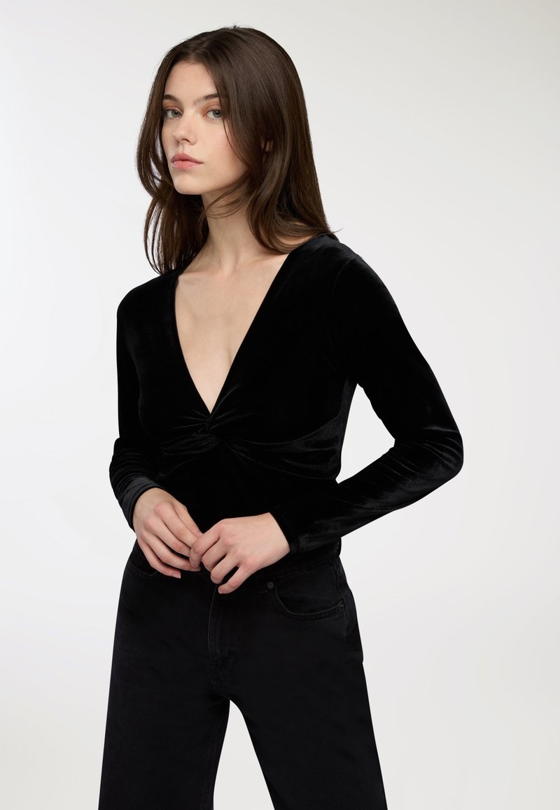 Młoda kobieta z długimi brązowymi włosami, ubrana w czarną aksamitną bluzkę z długim rękawem i głębokim dekoltem w kształcie V oraz czarne spodnie, stojąca na tle białym.