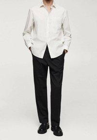 Personne portant une chemise blanche à manches longues rayée avec des boutons, un pantalon noir et des chaussures noires, debout les mains dans les poches.