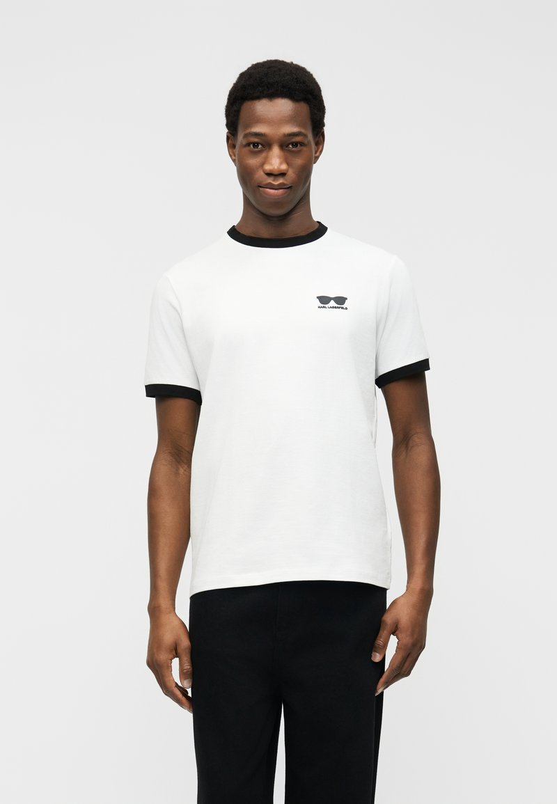 Νεαρός άνδρας που φοράει λευκό μπλουζάκι με μαύρο γιακά και μανσέτες και μικρό μαύρο λογότυπο γυαλιών ηλίου στο αριστερό στήθος, συνδυασμένο με μαύρο παντελόνι.