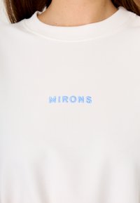 Witte sweatshirt met lichtblauwe geborduurde tekst "MIRONS." Gemaakt van zachte stof met een ronde halslijn en een gladde textuur.