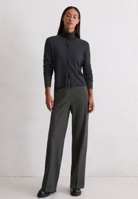 Sieviete stāv pie baltās sienas, uzvilkusi melnu pogātu kardigānu pār tumšo augsto kaklu, pelēkas platās bikses un melnas kurpes.