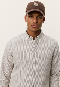 Καφέ καπέλο μπέιζμπολ με κεντημένο λογότυπο, συνδυασμένο με πουκάμισο με λεπτές κάθετες ρίγες σε ανοιχτό και σκούρο χρώμα.