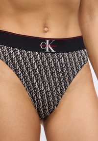 Μαύρα εσώρουχα με λευκό μοτίβο ολόκληρου του υφάσματος με το λογότυπο "CK", με αντίθετη μαύρη ζώνη μέσης που περιλαμβάνει κόκκινες λεπτομέρειες και εμπορική σήμανση.