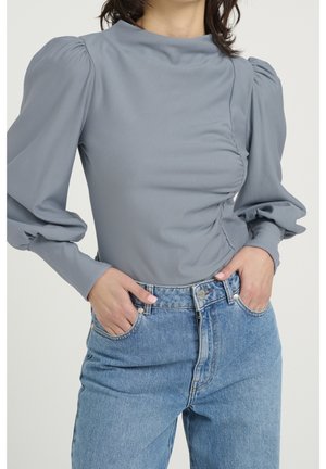 Femme portant une blouse grise à manches longues avec des épaules bouffantes et un jean bleu taille haute, les mains reposant dans les poches avant.
