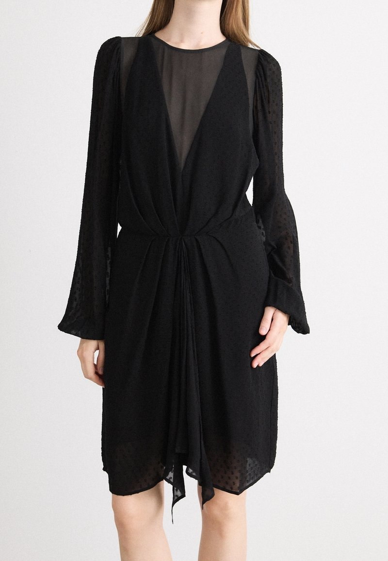Robe noire longueur genou avec manches longues en tulle à pois, un panneau transparent en V profond et un détail de tissu froncé à la taille.
