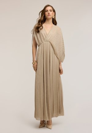 Frau steht und trägt ein beigefarbenes plissiertes Maxikleid mit einem asymmetrischen Ärmel, goldene Ohrringe, Armband und beigefarbene spitze High Heels.