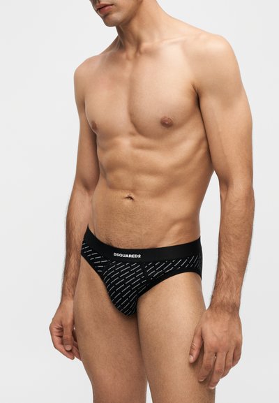 Modèle masculin portant un slip noir de marque DSQUARED2, debout devant un fond clair uni, visible du cou jusqu'aux cuisses.