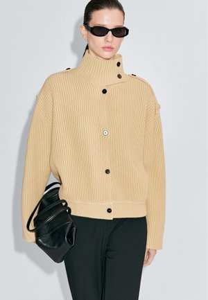 Maglione beige a coste con colletto alto, dotato di bottoni neri e una vestibilità rilassata. Abbinato a una borsa nera e pantaloni scuri, su uno sfondo neutro.