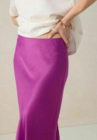 Jupe maxi en satin de couleur magenta vif, avec une texture lisse et une taille cintrée. À porter avec un haut blanc légèrement rentré.