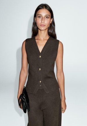 Braune, ärmellose Weste mit V-Ausschnitt, mit goldenen Knöpfen. Kombiniert mit passenden Hosen, dazu eine schwarze Clutch. Glatte Textur.