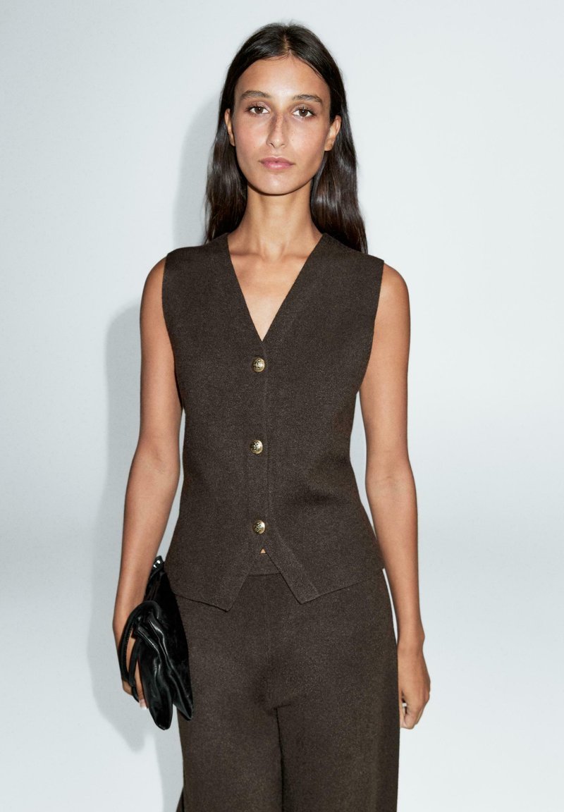 Veste marron sans manches à col en V, ornée de boutons dorés. Portée avec un pantalon assorti, accompagnée d'une pochette noire. Texture lisse.