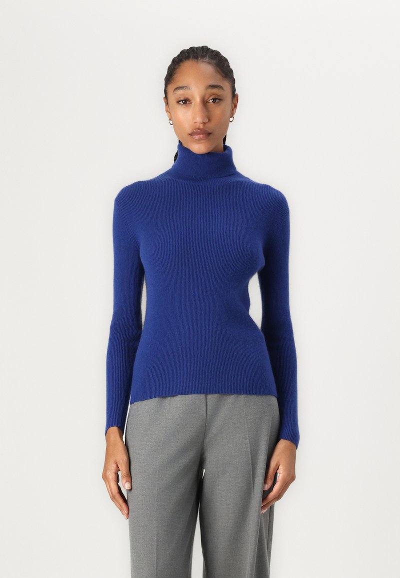 Suéter de cuello alto azul royal con textura de canalé, ajuste slim, mangas largas y un escote alto, combinado con pantalones gris ajustados.