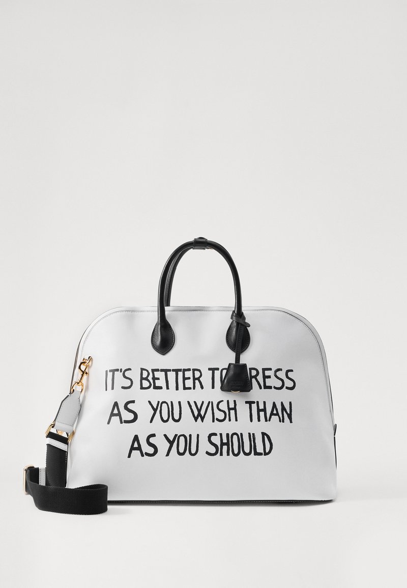 Hvid lædertaske med sorte accenter, der har et top håndtag og en aftagelig rem. Teksten trykt på forsiden lyder: "DET ER BEDRE AT KLÆDE SIG SOM DU VIL END SOM DU BURDE."