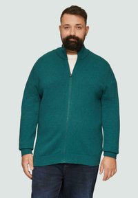 Tealfarbener Reißverschluss-Pullover aus Strickstoff, mit hohem Kragen und gerippten Bündchen, über einem weißen T-Shirt getragen, vor einem hellen Hintergrund.