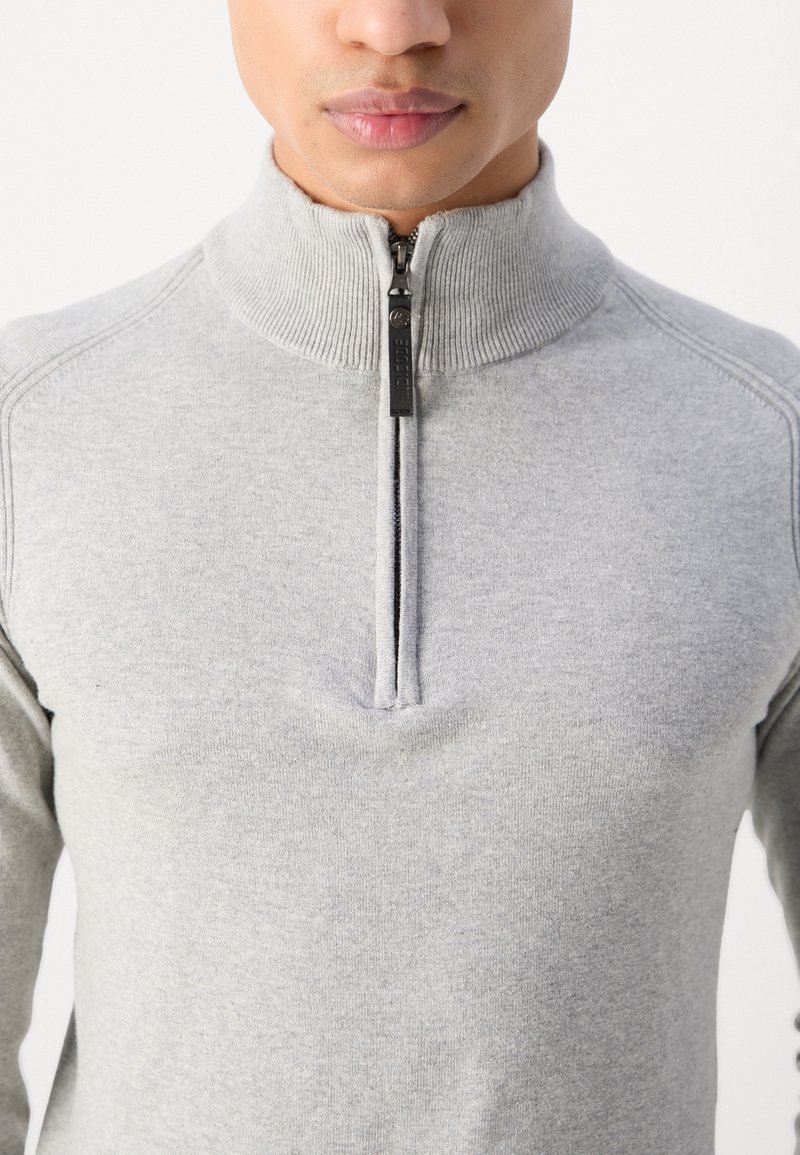 Maglione grigio chiaro con zipper e colletto alto. Presenta polsini e scollatura a coste. Texture morbida con un design aderente, dettagli di cucitura minimi.