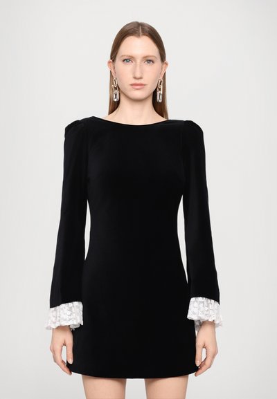 Robe en velours noir à manches longues, ornée de accents en dentelle blanche au niveau des poignets. Silhouette droite avec un col en bateau. Texture lisse.