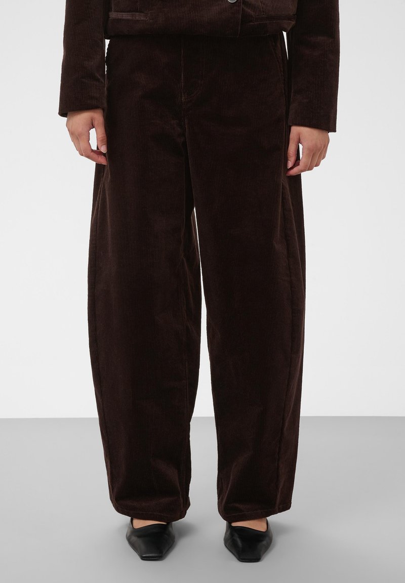 Personne portant un pantalon large en velours côtelé marron foncé et une veste assortie, debout les mains détendues le long du corps sur un fond uni.