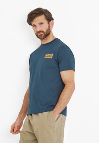 Man with beard wearing a navy blue t-shirt with "Endless Summer" text and beige pants, standing with one hand in pocket against white background.