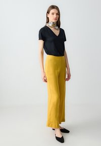 Zwarte V-hals T-shirt gecombineerd met hooggewaisted, brede mosterdgele broek, aangevuld met een lichte gestreepte sjaal en zwarte instapschoenen.
