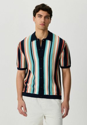 Jonge man die een kort-sleeve, knoopsluitend gebreid shirt draagt met verticale strepen in marineblauw, lichtblauw, beige en oranje, gecombineerd met een witte broek.
