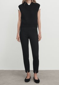 Haut en tricot noir sans manches avec un décolleté en V et un ourlet côtelé, associé à un pantalon noir ajusté avec des fentes latérales aux chevilles et des chaussures plates.