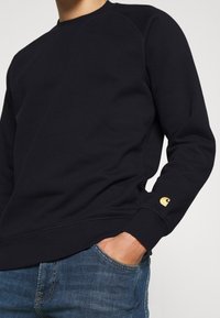 Navy sweatshirt made of cotton blend, featuring a crew neck, ribbed cuffs, and a gold logo accent on the left sleeve. Shown with denim jeans.