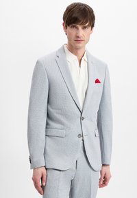 Hellgraue Anzugjacke mit strukturierter Oberfläche, Reverskragen, zwei Fronttaschen und einem roten Einstecktuch. Kombiniert mit einem cremefarbenen Hemd mit Kragen.