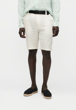 Mężczyzna ubrany w jasnobeżowe szorty, jasnooliwianą koszulę z długim rękawem włożoną w spodnie, czarny pleciony pasek i czarne espadryle wsuwane na stopę, stojący na białym tle.