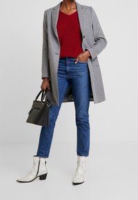 Grijze lange jas over een rode geribbelde trui, blauwe skinny jeans, witte enkellaarsjes met slangenprint en een zwarte handtas met handvatten.