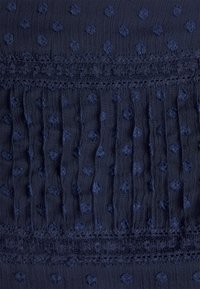 Tissu en bleu marine texturé présentant des pois en relief, du panneau et une dentelle en bordure, créant une apparence superposée et structurée.