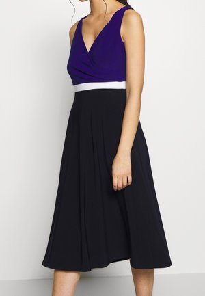 Kobieta w sukience bez rękawów z fioletowym dopasowanym topem, białym paskiem w talii i czarną plisowaną spódnicą midi na jednolitym tle.