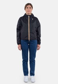 Giacca nera in nylon con cappuccio, dotata di una zip centrale con dettaglio giallo, indossata sopra a jeans blu e sneakers bianche.