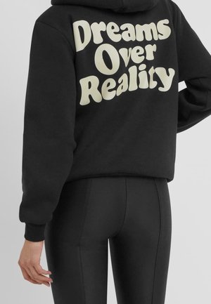 Persoon draagt een zwarte hoodie met grote crèmekleurige tekst "Dreams Over Reality" op de rug en zwarte aansluitende broek, hand ontspannen langs het lichaam.