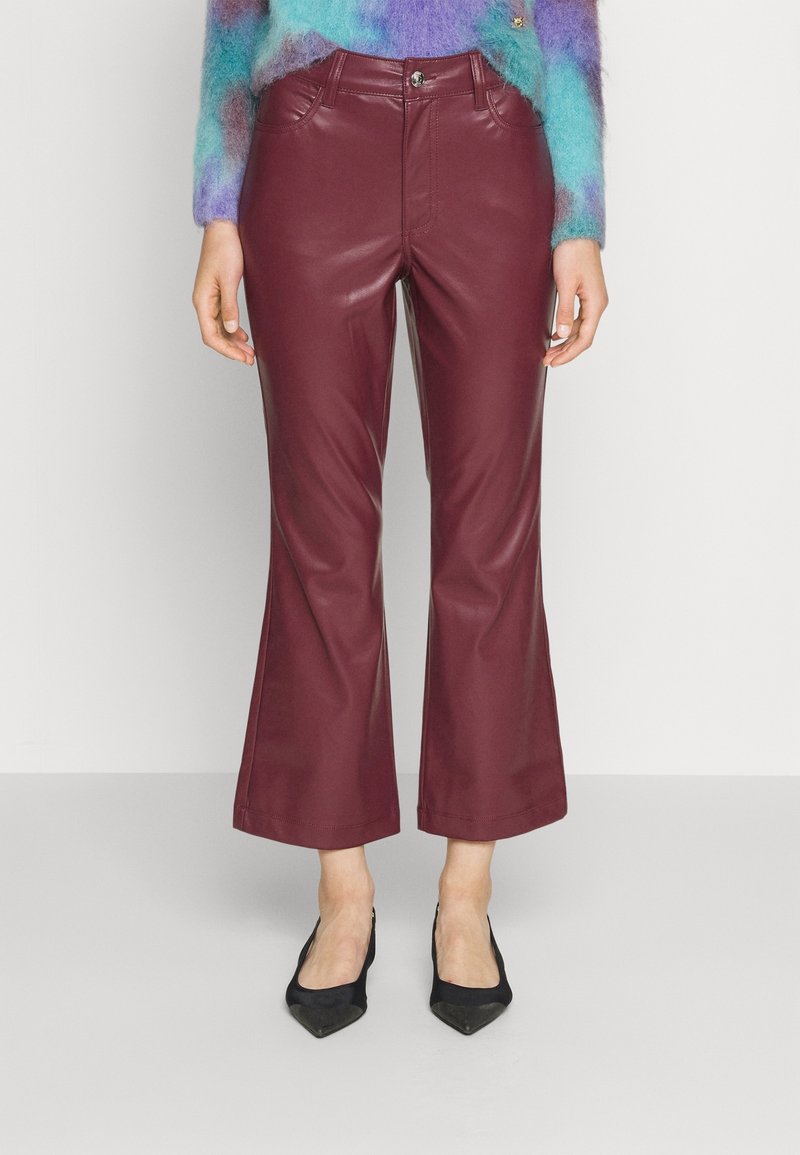 Femme portant un pantalon en cuir bordeaux taille haute, coupe flare courte avec poches avant, associé à des talons noirs à bout pointu et un pull multicolore en matière douce.