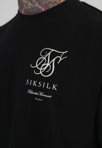 Camiseta de algodón negra con cuello redondo. Presenta un logo en blanco y el texto "SIKSILK" encima de "Sigue el movimiento" y "Est 2012" en el dobladillo.