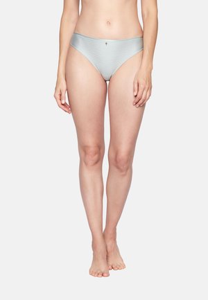 Frau trägt silberfarbene Bikini-Unterwäsche mit Streifen, steht barfuß vor einem schlichten hellgrauen Hintergrund.