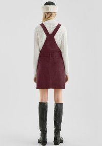 Burgunderfarbene Corduroy-Latzkleid mit verstellbaren Trägern, das eine figurschmeichelnde Silhouette bietet, Taschen hat und mit schwarzen Leder-Stiefeln bis zum Knie kombiniert ist.
