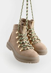 Stivaletti alla caviglia in suede beige con accessori dorati e lacci a motivo verde-nero. Suola in gomma testurizzata e colletto imbottito per il comfort.