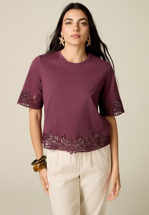 Kvinde iført en lilla top med blondekant på ærmer og kant, beige bukser, søstjerneøreringe og brune og sorte armbånd på venstre håndled.