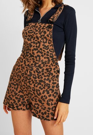 Kvinde iført brune leopardmønstrede korte overaller med sorte knapper over en langærmet sort lynlås-top mod en ensfarvet baggrund.