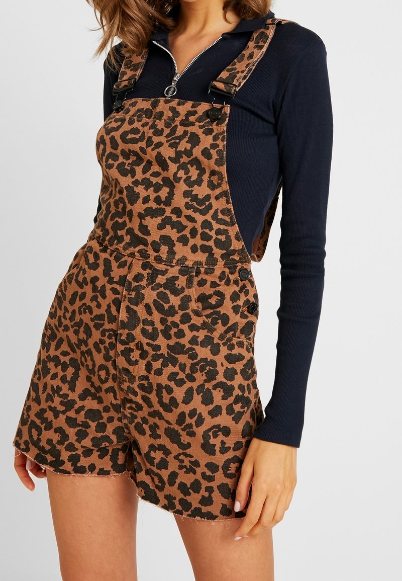 Femme portant une salopette courte imprimée léopard marron avec des boutons noirs, par-dessus un haut noir zippé à manches longues, sur un fond uni.