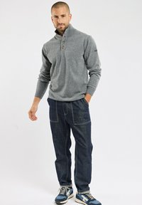 Pull en tricot gris avec un col haut et une patte de boutonnage à trois boutons, associé à un pantalon en denim bleu foncé avec des détails de couture visibles et des poches.