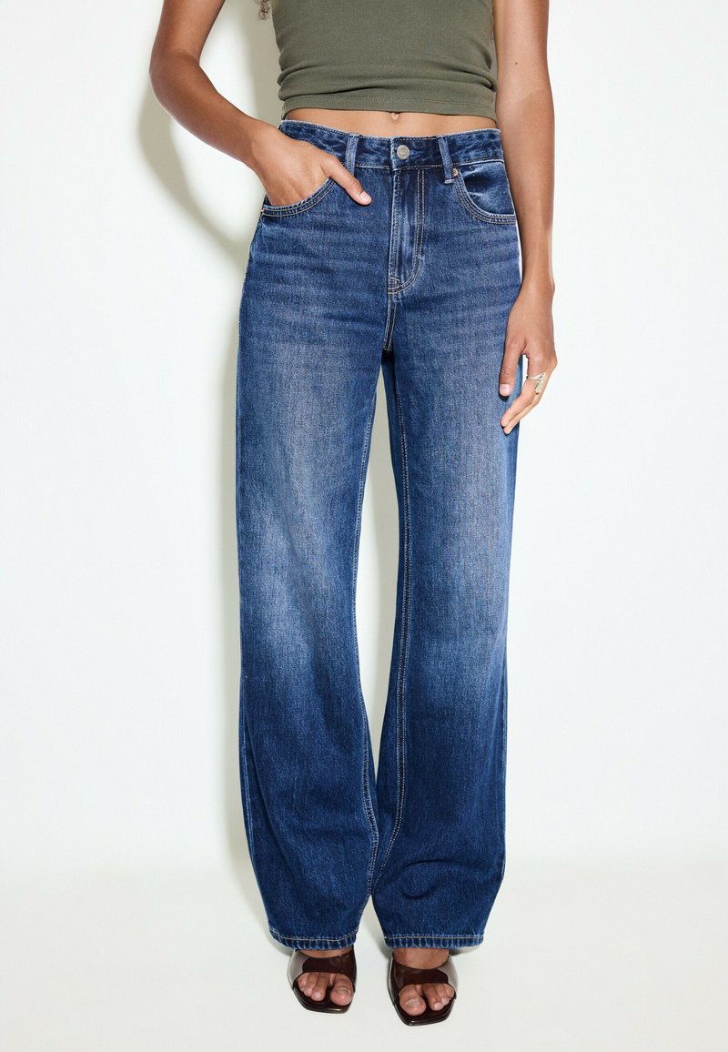 Femme portant un jean bleu taille haute à jambes larges, un débardeur court vert olive et des talons ouverts marron, debout sur fond blanc.