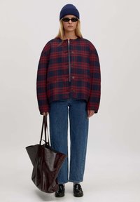 Rood en marineblauw geruite oversized jas met ronde halslijn, zes knopen en twee zijzakken; gecombineerd met blauwe jeans en een donkere draagtas.