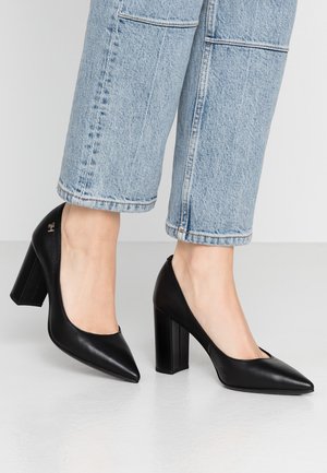 Sorte læderstiletter med spids tå og blokhæl. Glat tekstur med en diskret mærkeaccent på hælen. Kombineret med afklippede blå jeans.