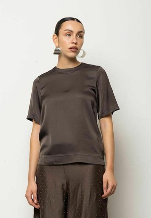 Donna con capelli raccolti all’indietro, che indossa grandi orecchini a cerchio argentati, una maglietta corta marrone scuro lucida e pantaloni a pois abbinati.