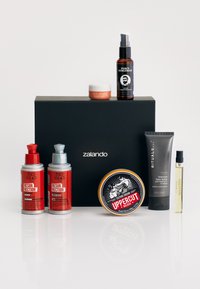 Osiem produktów do pielęgnacji uporządkowanych przed czarnym pudełkiem, w różnych kolorach i wzorach: dwie czerwone butelki, brązowy olejek, szary tubka i złota puszka.