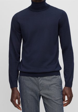 Homme portant un pull à col roulé bleu marine et un pantalon gris texturé sur un fond uni.